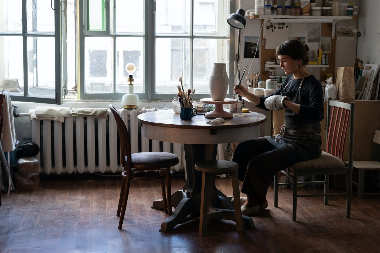 young woman in apron sits on chair at round table painting handmade ceramic vase in workshop.jpg